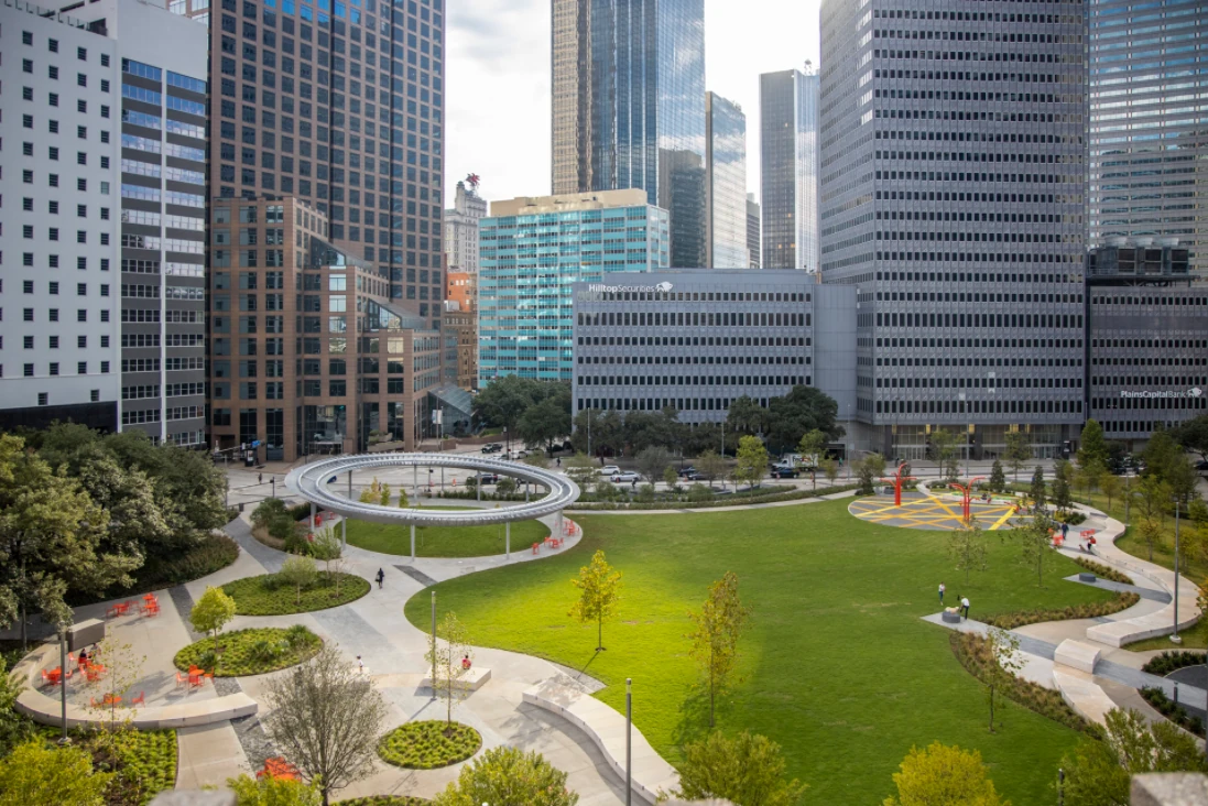 Pacific Plaza from above, among downtown Dallas towers: the ring-shaped pavilion, the lawn, a yellow-striped plaza and curving paths